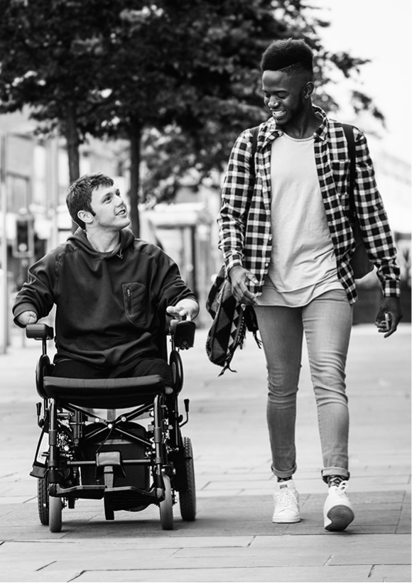 Foto preto e branca mostra dois jovens na rua. Um deles possui deficiência e usa cadeira de rodas.